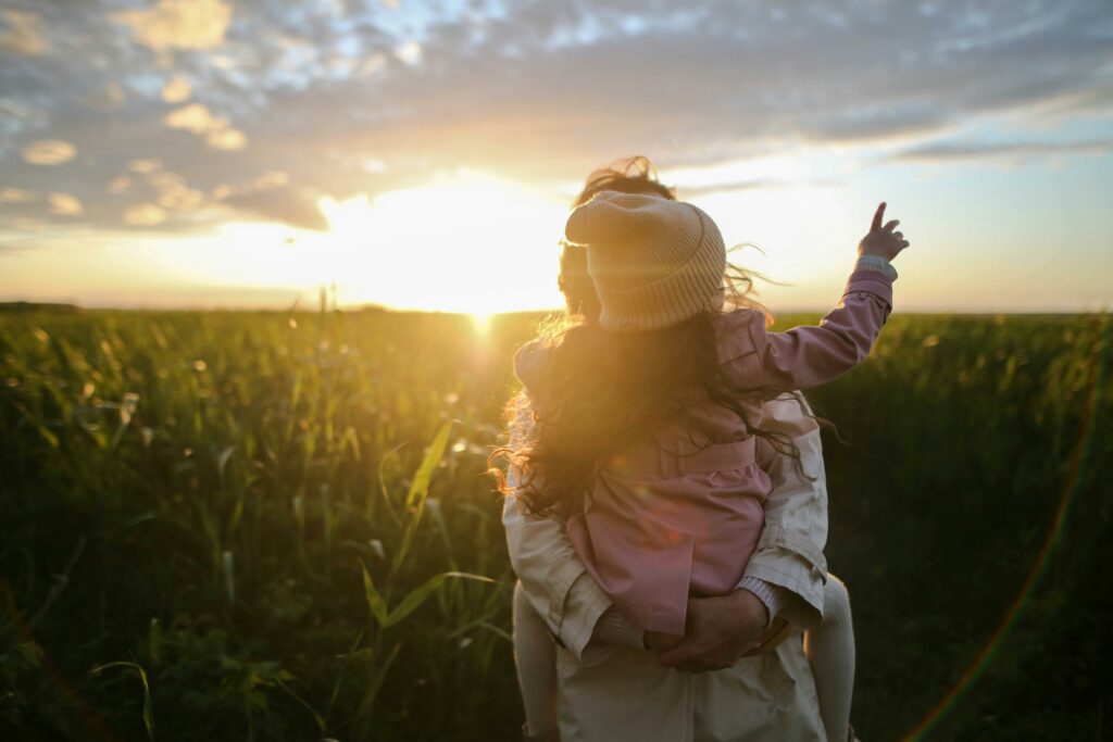 pexels-photo-1683975-1683975 A mother and daughter embrace and point at the sunset in a grassy field.